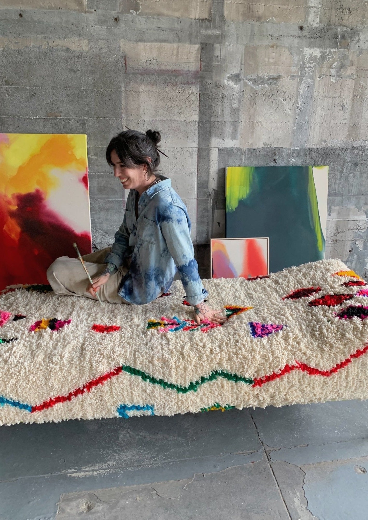 artist sits on chaise lounge day bed holding her paint brush in between paintings. She is able to rest contemplate and regain vision in her artwork while staying inspired inside her urban art studio.